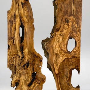 May include: Two vertical, irregular-shaped wooden sculptures with a natural, organic aesthetic. The wood grain is prominent, with swirling patterns in shades of brown and tan. Each piece has a large, irregular hole. The overall effect is rustic and artistic.