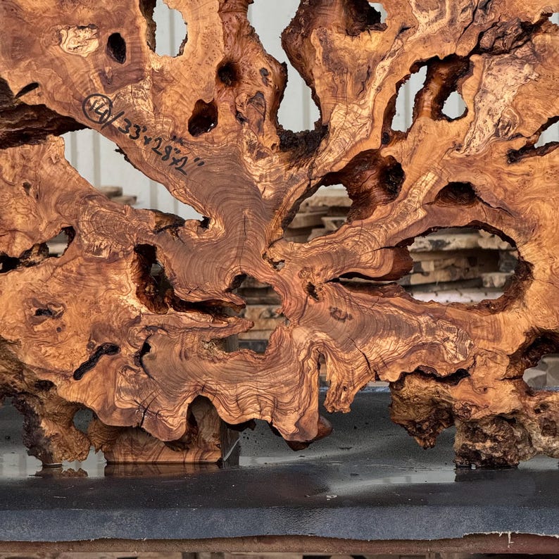 May include: A natural olive wood slab featuring a complex, swirling grain pattern with multiple organic voids and irregular edges. The wood surface displays a warm brown color with darker streaks and includes handwritten text reading 40 x 33 x 28 1/2 inches.