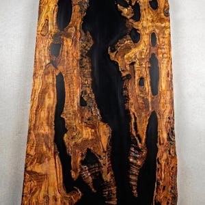 May include: A large, rectangular wooden slab with a striking design. The wood features a natural, warm brown color with intricate grain patterns and dark, irregular voids. A central, glossy black resin fills the gaps, creating a dramatic contrast.