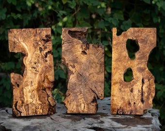 Könnte beinhalten: Drei aufrecht stehende, rechteckige Holzplatten mit komplexen natürlichen Maserungen, Astlöchern und unregelmäßigen Kanten. Das Holz zeigt eine satte goldbraune Farbe mit dunklen, wirbelnden Texturen und organischen Hohlräumen.