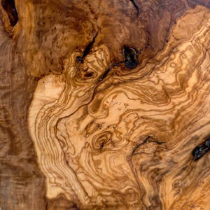 May include: A close-up view of a polished olive wood surface featuring intricate, swirling grain patterns in shades of light tan, golden yellow, and deep brown. The wood displays natural knots, dark organic fissures, and a smooth texture.