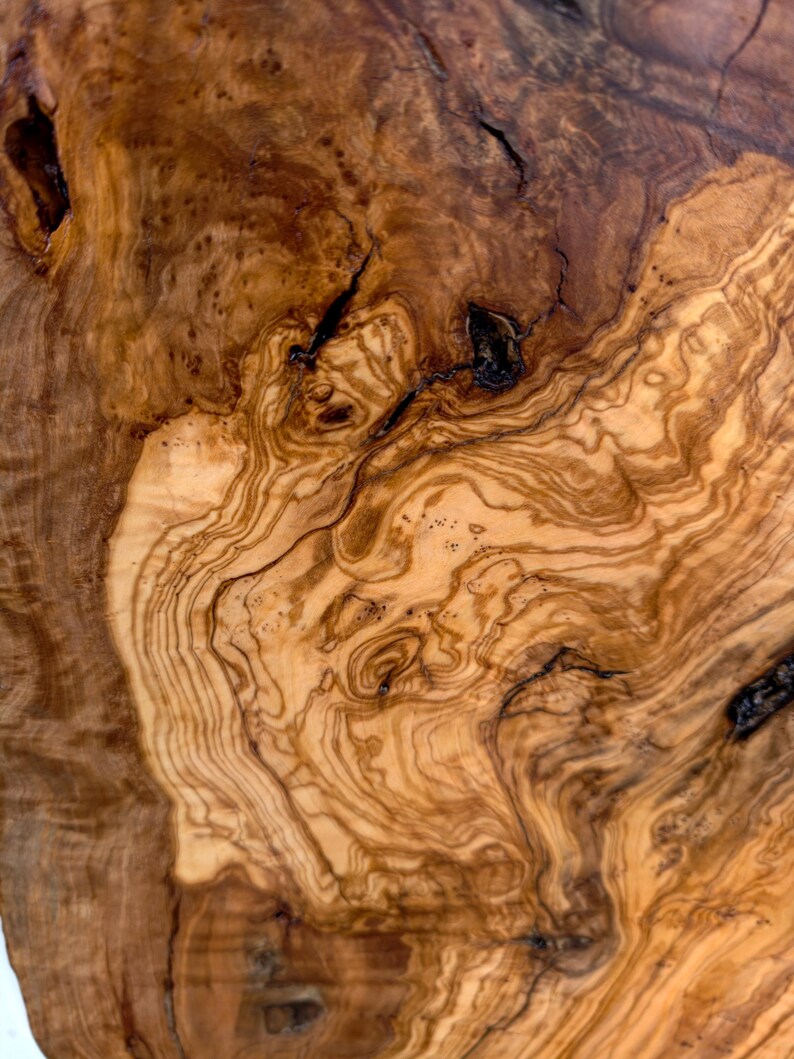 May include: A close-up view of a polished olive wood surface featuring intricate, swirling grain patterns in shades of light tan, golden yellow, and deep brown. The wood displays natural knots, dark organic fissures, and a smooth texture.