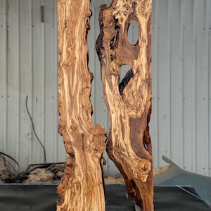 May include: Two vertical, raw-edge wooden slabs with a natural, organic aesthetic. The wood grain is prominent, showcasing a mix of light and dark brown tones. One slab has a large, irregular hole. The slabs are displayed against a neutral background.
