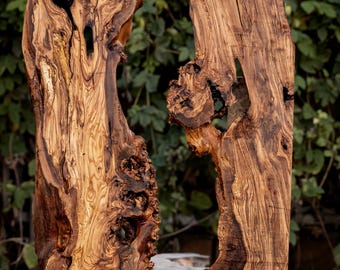 May include: Two vertical slabs of olive wood featuring prominent natural grain patterns, dark brown swirls, and irregular live edges. The wood surfaces display complex knots, organic textures, and deep color variations ranging from light tan to dark espresso.