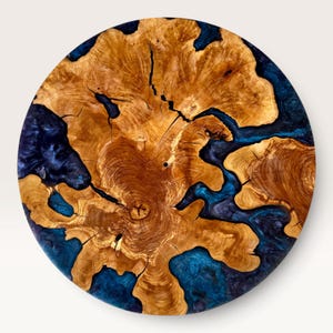May include: A circular tabletop featuring a natural light brown wood slab with organic edges, integrated with deep blue and translucent resin filling the gaps and cracks. The wood grain is visible, and the surface has a smooth, polished finish.