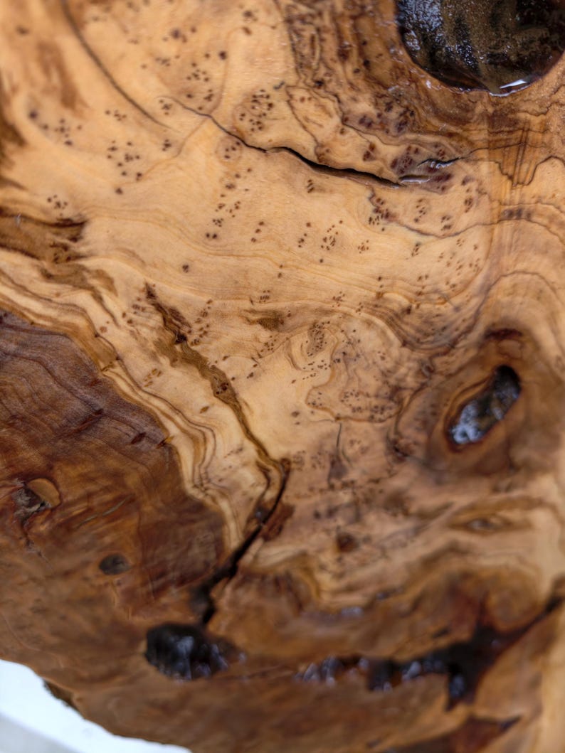 May include: A close-up view of natural olive wood grain featuring swirling patterns of light tan and deep brown tones. The surface displays organic knots, dark inclusions, and intricate, flowing lines that highlight the wood's unique texture.