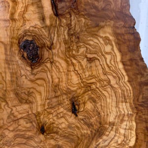 May include: A slab of olive wood featuring a complex, swirling grain pattern in shades of light tan, golden yellow, and deep brown. The surface displays several dark, prominent knots and organic, irregular edges that highlight the natural texture.