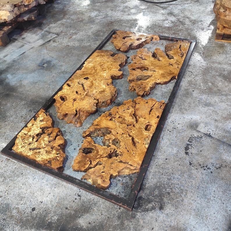 May include: A large wooden table top made of several pieces of wood with a natural wood grain pattern. The wood is a light brown color and has a lot of knots and burls. The table top is framed in a black metal frame.