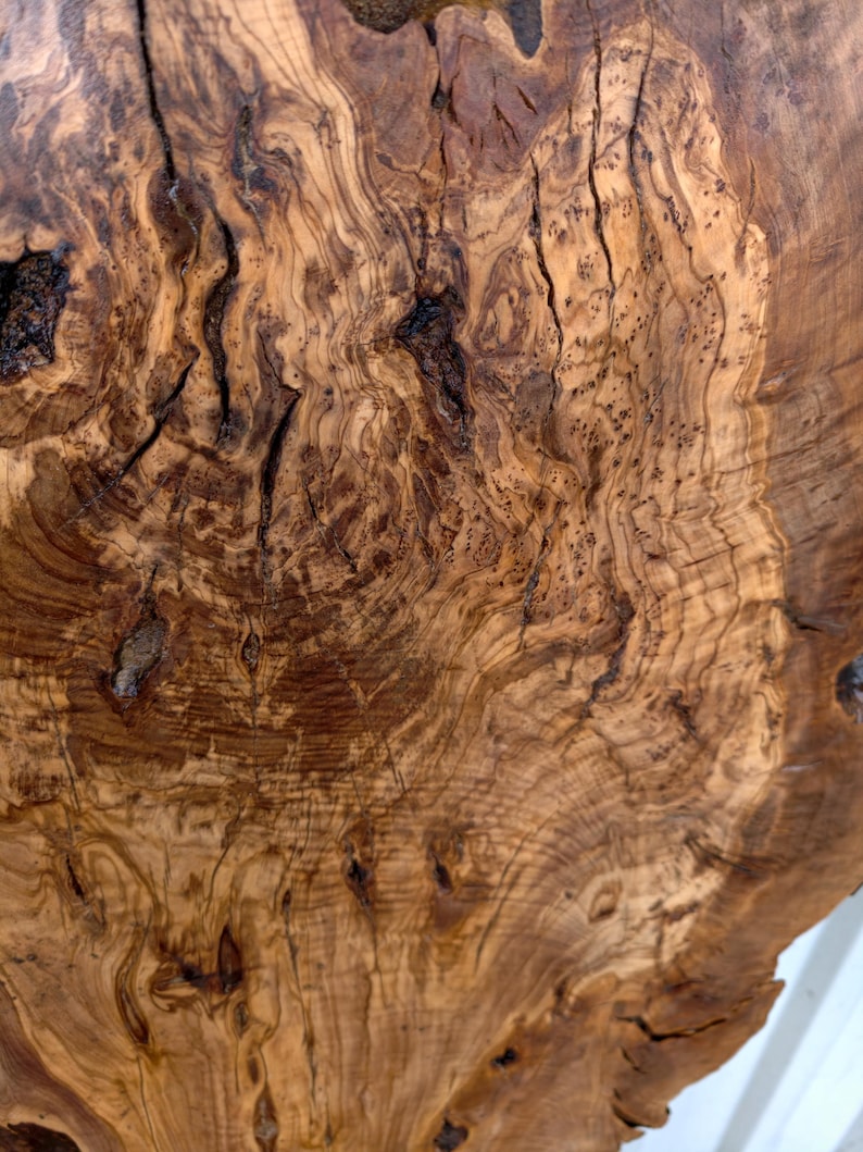 May include: A polished slab of olive wood featuring a complex, swirling grain pattern in shades of golden brown and dark chocolate. The surface displays natural knots, organic fissures, and intricate burl wood textures throughout the piece.