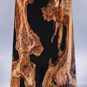 May include: A rectangular wooden table with a black epoxy resin center. The wood has a natural, organic shape with intricate grain patterns in shades of brown and tan. The table is approximately 30 inches long.
