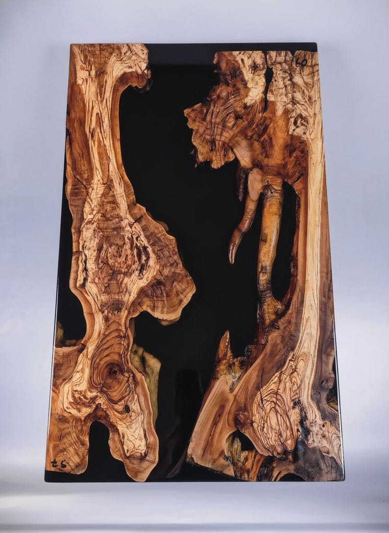 May include: A rectangular wooden table with a black epoxy resin center. The wood has a natural, organic shape with intricate grain patterns in shades of brown and tan. The table is approximately 30 inches long.