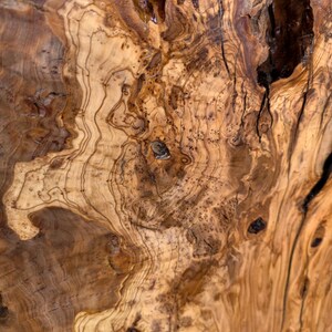 May include: A close-up view of a polished olive wood surface featuring intricate, swirling grain patterns in shades of light tan, golden yellow, and deep brown. The texture includes natural knots, organic voids, and dark, irregular streaks.