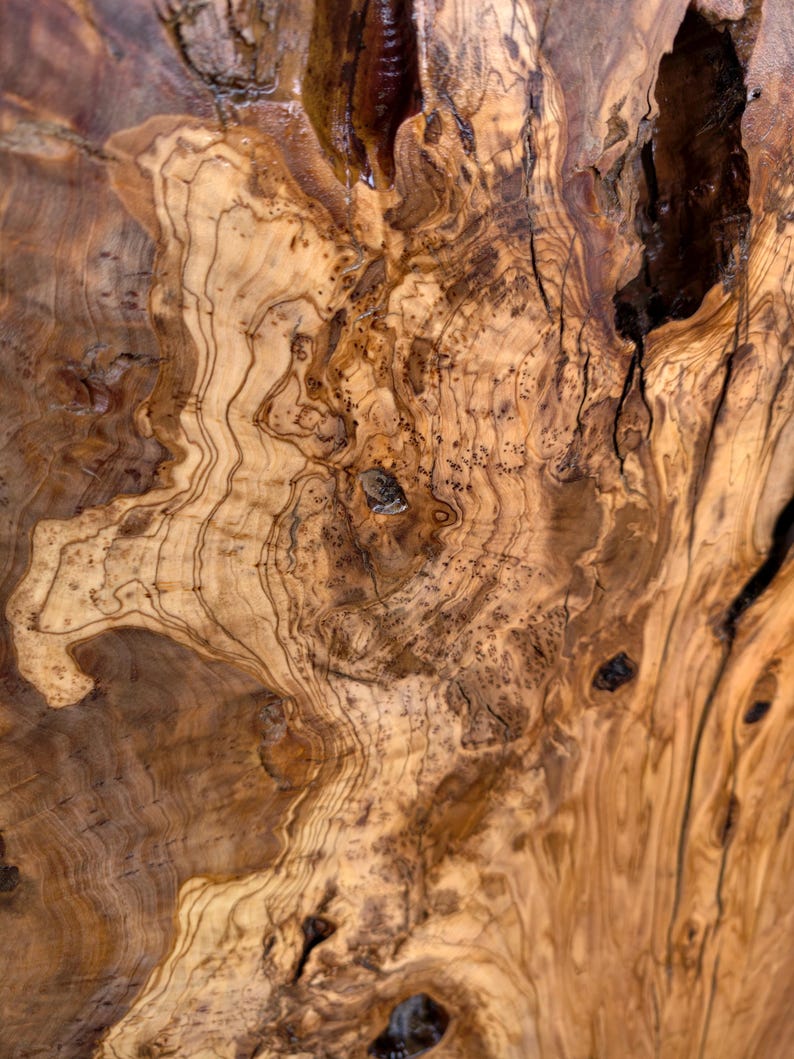 May include: A close-up view of a polished olive wood surface featuring intricate, swirling grain patterns in shades of light tan, golden yellow, and deep brown. The texture includes natural knots, organic voids, and dark, irregular streaks.