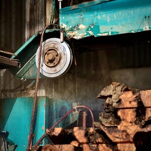 May include: A close-up shot of a circular saw blade in action, cutting through a piece of wood. The saw is part of a larger machine, with a teal-colored metal frame. Water sprays from the blade as it cuts. The wood is rough and brown.