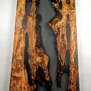 May include: A rectangular wooden table with a dark gray resin center. The wood has a natural, textured appearance with shades of brown and black. The table's design highlights the contrast between the wood and resin.
