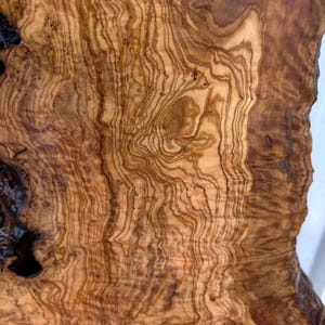 May include: A slab of olive wood featuring a complex, swirling grain pattern in shades of light tan, golden yellow, and deep dark brown. The surface displays natural organic curves, knots, and irregular edges with a smooth, polished finish.
