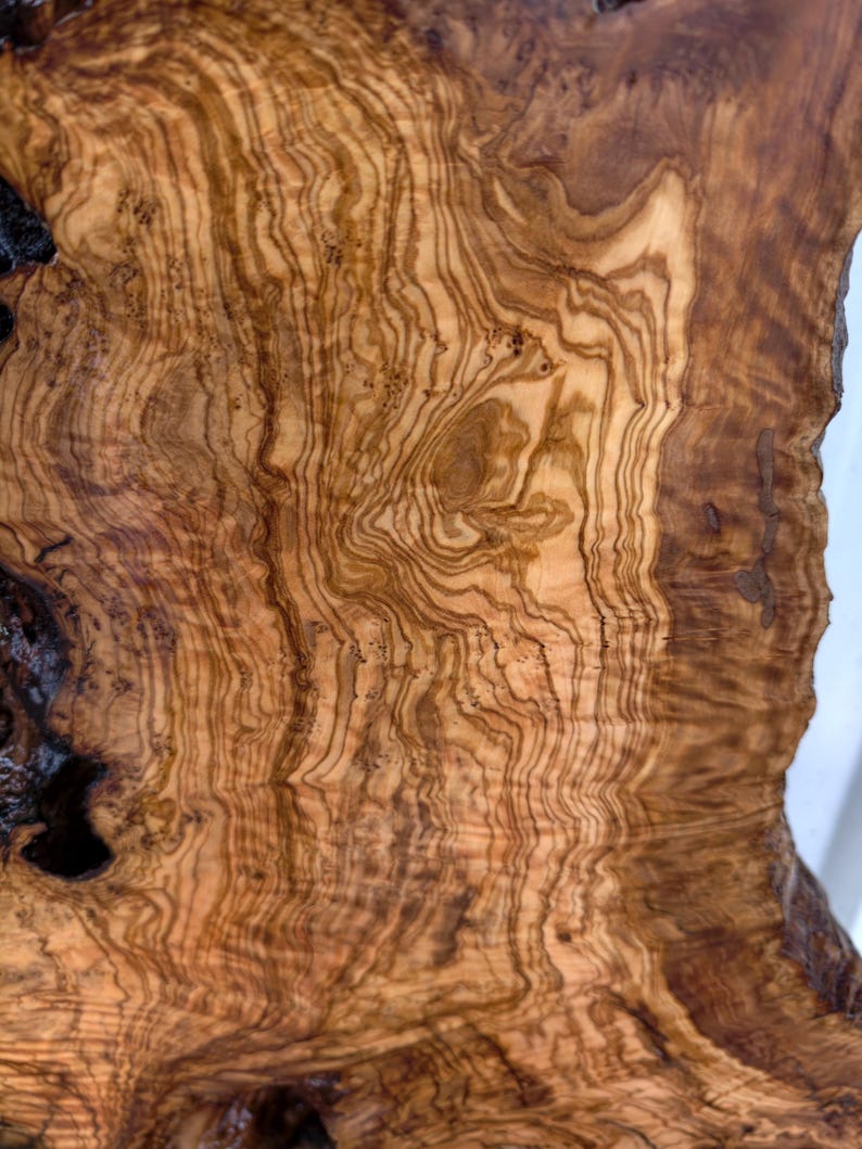 May include: A slab of olive wood featuring a complex, swirling grain pattern in shades of light tan, golden yellow, and deep dark brown. The surface displays natural organic curves, knots, and irregular edges with a smooth, polished finish.