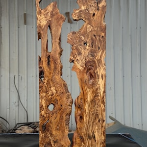 May include: Two vertical wooden slabs with a natural, organic shape. The wood has a rich, warm brown color with intricate grain patterns and knots. The slabs are roughly rectangular, with irregular edges and holes, suggesting a raw, unfinished aesthetic. The background is a neutral industrial setting.