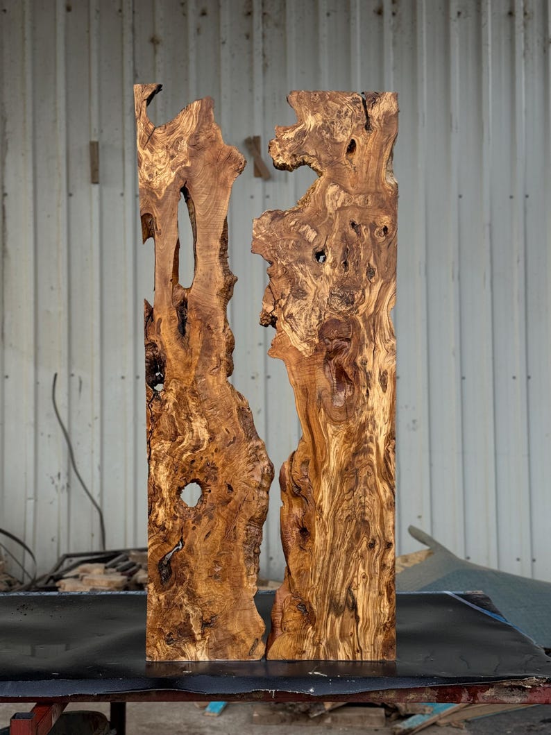 May include: Two vertical wooden slabs with a natural, organic shape. The wood has a rich, warm brown color with intricate grain patterns and knots. The slabs are roughly rectangular, with irregular edges and holes, suggesting a raw, unfinished aesthetic. The background is a neutral industrial setting.