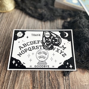Puede incluir: Una ilustración en blanco y negro de una tabla Ouija con las letras A-Z, los números 0-9 y las palabras "Thank You", "Goodbye" y "For Your Order". Una bola de cristal se encuentra en el centro de la tabla.