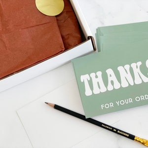 a flat lay image of the 2-inch round matte gold sticker being used in a shipping box to close the tissue wrap on an item ready to ship.   A pencil and thank you insert cards lay next to the box for context