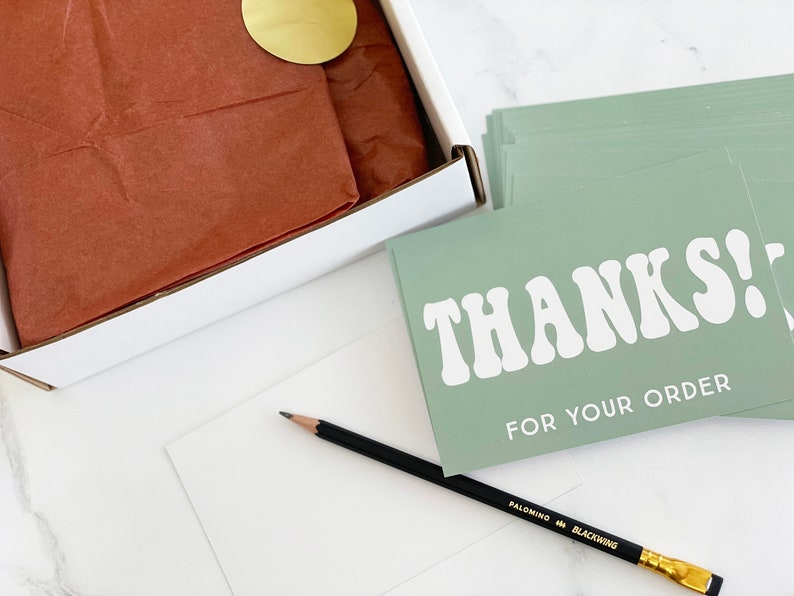 a flat lay image of the 2-inch round matte gold sticker being used in a shipping box to close the tissue wrap on an item ready to ship.   A pencil and thank you insert cards lay next to the box for context