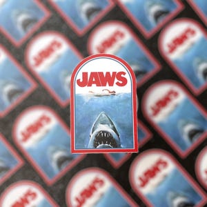 Puede incluir: Una pegatina roja y blanca con la palabra "JAWS" en letras blancas. La pegatina presenta un primer plano de la cabeza de un tiburón con la boca abierta, revelando dientes afilados. El tiburón nada en agua azul con un nadador en el fondo.