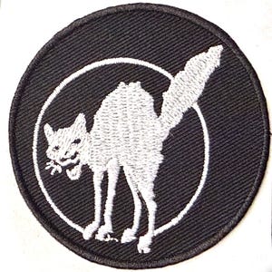 Op de afbeelding: Een ronde, zwarte patch met een wit geborduurd beeld van een kat in een witte cirkel. De kat is gebogen met zijn staart omhoog en mond open. De patch heeft een zwarte rand.