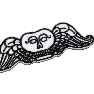 May include: An embroidered patch featuring a white skull with stylised black line details and large, sweeping white wings with black outlines. The design is symmetrical, with the wings extending outwards from the central skull shape on both sides.