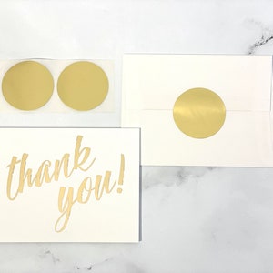 a flat lay view of a 2-inch round matte gold sticker shown in use on an A-4 envelope with a card and 2 unused stickers for perspective