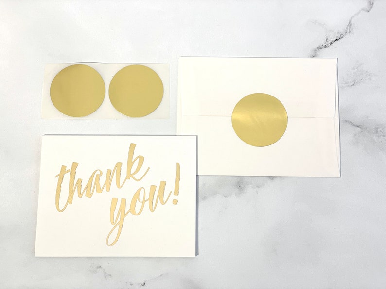 a flat lay view of a 2-inch round matte gold sticker shown in use on an A-4 envelope with a card and 2 unused stickers for perspective