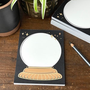 May include: A black notepad with a gold glitter design of a snow globe with stars. The snow globe is empty and ready for writing.