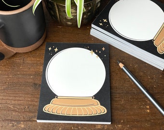 May include: A black notepad with a gold glitter design of a snow globe with stars. The snow globe is empty and ready for writing.