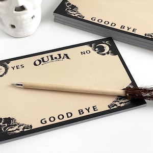 May include: A tan Ouija board with black borders and text, including "YES," "NO," and "GOOD BYE." A wooden pen with a tassel rests on the board. A stack of identical boards is visible.