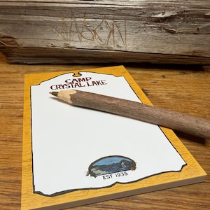 May include: A rectangular notepad with a yellow border and white centre featuring the text Camp Crystal Lake and a small lake illustration with the text EST 1935. A rustic wooden twig pencil with a sharpened graphite tip rests on the notepad.