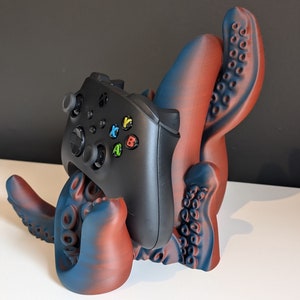 Könnte beinhalten: Ein 3D-gedruckter, krakenförmiger Controller-Ständer mit einem rot-blauen Farbverlauf. Der Ständer hält einen schwarzen Xbox-Controller.