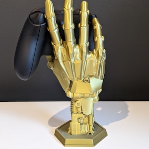 May include: A gold-colored, 3D printed, robotic hand holding a black video game controller. The hand is attached to a base with a hexagonal shape.