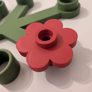 Peut inclure: Un objet rouge en forme de fleur avec un trou central, ainsi que des éléments verts en forme de tige et d'anneau. Les objets semblent être imprimés en 3D. La fleur a un design de pétales et est le point central de l'image.