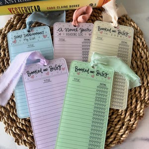 May include: Five rectangular reading log bookmarks in light blue, pink, cream, lavender, and light green, each featuring a ribbon at the top. The bookmarks include printed text such as Shelf Improvement, A Novel Year, and Booked and Busy, with lines for book titles and heart-shaped rating icons.