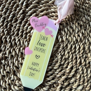 May include: A pencil-shaped bookmark with the words "Teach Love Inspire" and "Happy Valentine's Day!" printed on it. The bookmark has a pink ribbon and two pink heart accents. The top of the bookmark says "Miss Buckley".