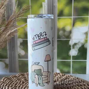 May include: A white stainless steel tumbler with a clear straw. The tumbler features illustrations of books, a cozy armchair, and the word "TBR2" and "Cozy". Pink and light blue hearts and crosses are scattered around the design.
