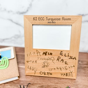 May include: A wooden picture frame with a white mat and a clear glass front. The frame is engraved with the text "KI ECC Turquoise Room 2022-2023". The bottom of the frame has a handwritten message with the names of children.