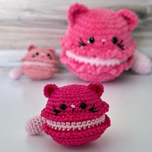 May include: Three crocheted cat-shaped macaron toys in shades of pink and fuchsia. Each features small ears, embroidered black eyes, whiskers, and a light pink filling layer. The text "hooks and scraps" is printed in black at the bottom of the image.