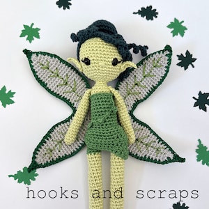 Può includere: Una bambola all'uncinetto con pelle verde chiaro, capelli verde scuro e un vestito verde. Presenta grandi ali a forma di foglia con centri grigi e bordi verde scuro. Il testo "hooks and scraps" è stampato in lettere nere sotto la bambola.