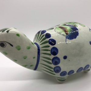 May include: Hand-painted ceramic turtle figurine with green, blue and white accents. The turtle has a detailed shell with a floral design and a whimsical face.