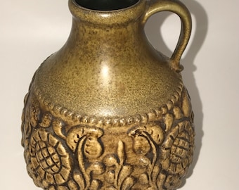 May include: A brown ceramic vase with a textured floral design. The vase has a wide base and a narrow neck with a handle.