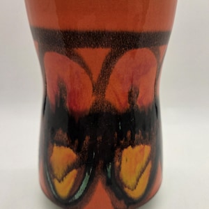 Könnte beinhalten: Eine Keramikvase mit ausgestelltem Rand und geschwungenem Körper. Die Vase hat einen leuchtend orangefarbenen Hintergrund mit einem abstrakten Design in Schwarz, Rot und Gelb. Der Rand ist weiß.