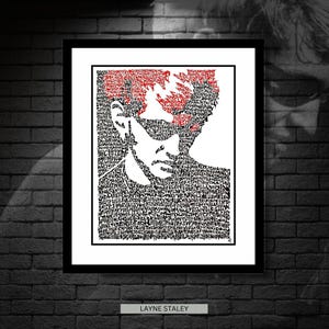 May include: A framed black and white portrait of Layne Staley, with red hair, created from text. The name "LAYNE STALEY" is printed in white on a black rectangle at the bottom. The artwork is displayed on a dark brick wall.