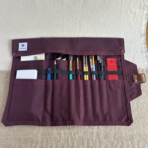 May include: A burgundy canvas roll-up case with a black elastic band and multiple slots for holding paintbrushes and pencils. The case has a label that says "Sal Original".