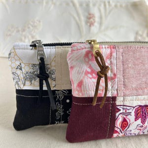 Puede incluir: Dos bolsas de cremallera de tela patchwork con tiradores de cremallera de cuero marrón. Una bolsa tiene un patrón negro, blanco y dorado, y la otra tiene un patrón rosa, blanco y burdeos.